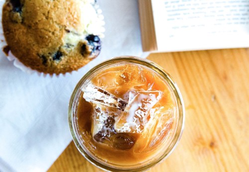 A top view of a glass of iced coffee with ice cubes, next to a blueberry muffin and an open book on a wooden table.