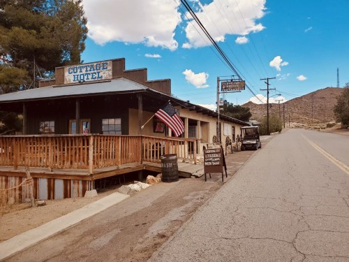 A rustic hotel with a wooden porch, American flag, and signs, set against a clear blue sky and mountains in the background.