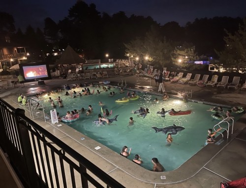 A lively nighttime pool scene with children and adults swimming, floating on inflatables, and watching a movie on a screen.