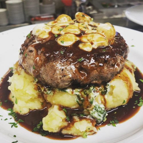 A juicy hamburger patty topped with gravy, served on a bed of creamy mashed potatoes and garnished with herbs.
