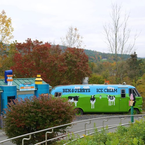 A colorful Ben & Jerry's ice cream truck parked near a building, surrounded by autumn foliage and trees.