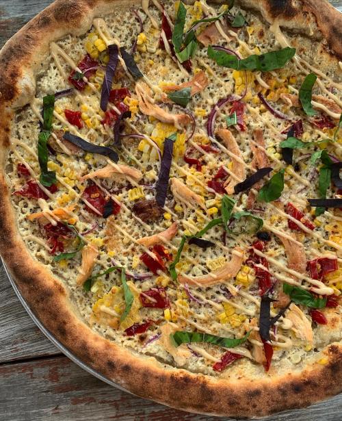 A close-up of a pizza topped with chicken, corn, red onions, basil, and drizzled with sauce on a golden crust.