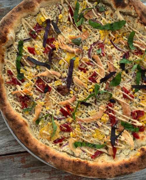 A close-up of a pizza topped with chicken, corn, red onions, basil, and drizzled with sauce on a golden crust.