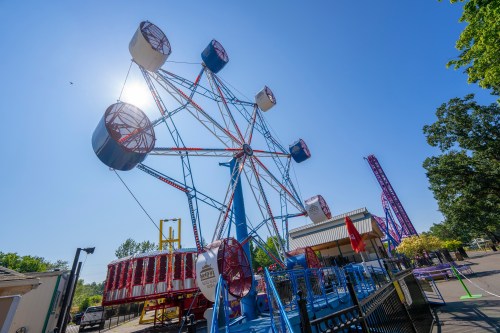 A colorful Ferris wheel stands against a clear blue sky at an amusement park, with rides and trees in the background.