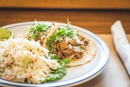 Two tacos filled with shredded meat, topped with onions and greens, served with a side of coleslaw and a lime wedge.