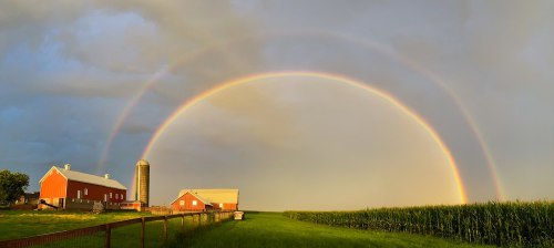 A vibrant double rainbow arcs over a red barn and silo, with green fields and corn in the foreground.