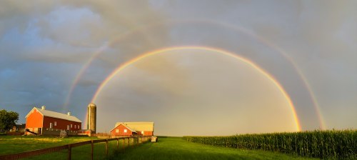 A vibrant double rainbow arcs over a red barn and silo, with lush green fields and a cloudy sky in the background.