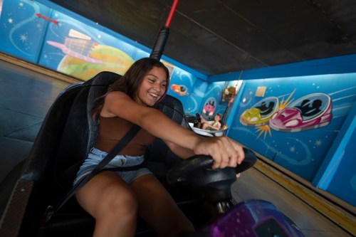 A young girl joyfully drives a bumper car, smiling as she grips the steering wheel in a colorful amusement park setting.
