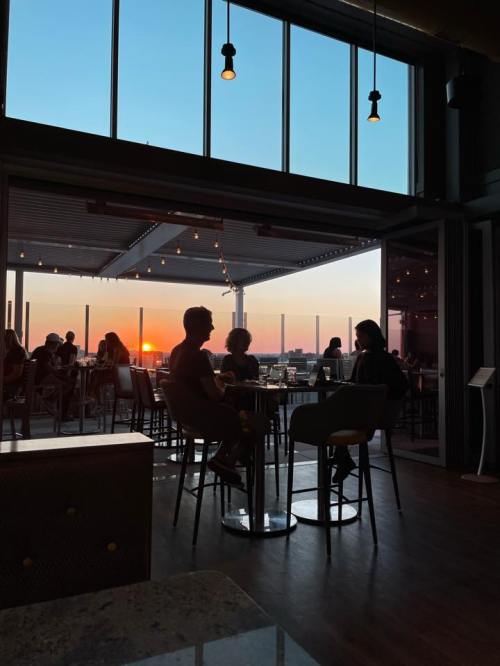 Silhouettes of people sitting at a bar, with a sunset view through large windows and warm lighting.