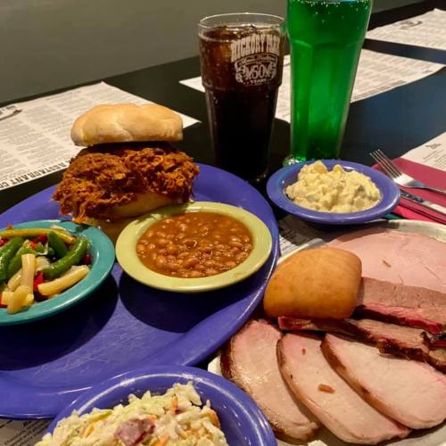 A colorful plate of barbecue with pulled pork sandwich, sliced ham, baked beans, coleslaw, and green salad, alongside drinks.
