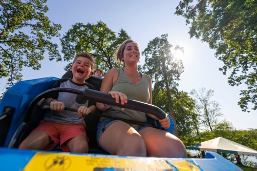A child and a woman enjoy a thrilling ride at an amusement park, smiling under a sunny sky with trees in the background.