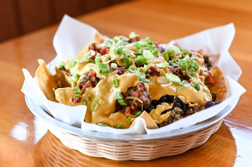 A basket of nachos topped with cheese, black beans, diced tomatoes, and green onions on a wooden table.