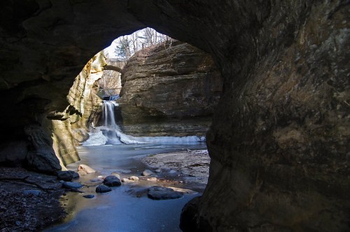 A serene view of a waterfall cascading through a rocky archway, surrounded by ice and natural rock formations.
