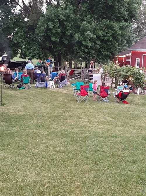 A gathering of people sitting in lawn chairs on a grassy area, with trees and a red barn in the background.