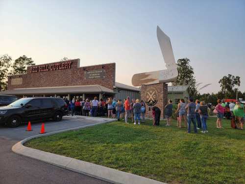 A long line of people waiting outside Red Hill Cutlery, with a large knife sign and parked police vehicles nearby.