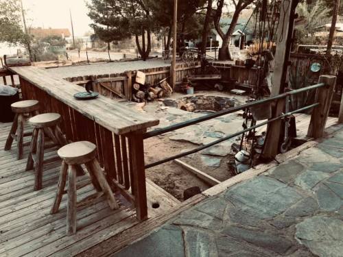 A rustic outdoor space with a wooden bar, stools, and a stone area surrounded by trees and a fence.
