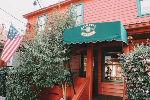 Exterior of Gold Dust Pizza, a red building with a green awning, surrounded by greenery and an American flag.
