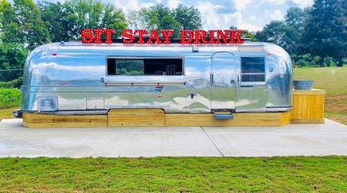 A shiny silver Airstream trailer with a wooden deck, featuring a neon sign that says "SIT STAY DRINK."