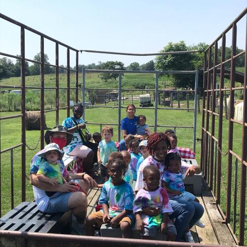 A group of children and adults in tie-dye shirts sitting in a fenced area outdoors, enjoying a sunny day.