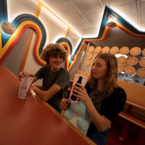 Two young people sit together in a colorful setting, enjoying drinks and smiling at each other.
