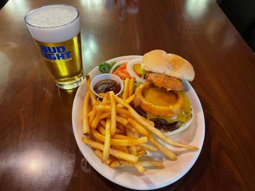A plate with a burger topped with pineapple, fries, a small bowl of sauce, and a glass of beer.