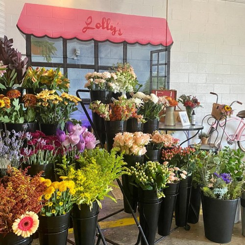 A vibrant flower shop display with various colorful blooms in black buckets, set against a pink awning backdrop.
