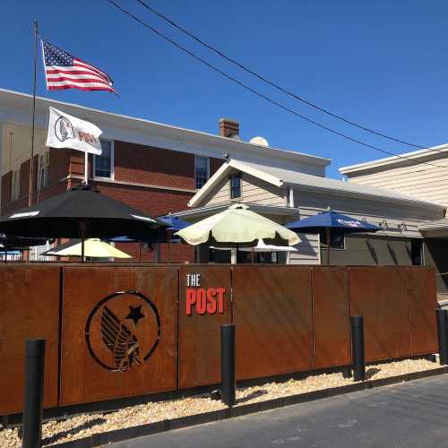 Outdoor seating area at The Post, featuring umbrellas and flags, with a rustic metal fence and a building in the background.