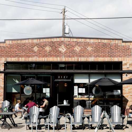 A brick building with large windows and outdoor seating, featuring umbrellas and patrons enjoying their time.