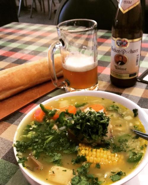 A bowl of soup with vegetables and herbs, a beer, and a breadstick on a checkered tablecloth.