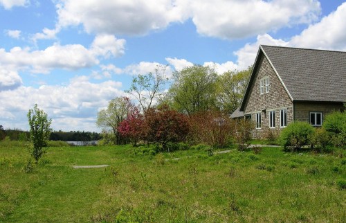 A serene landscape featuring a house beside a lush green field, trees, and a calm body of water under a cloudy sky.