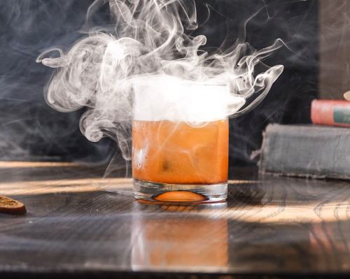 A glass of smoky cocktail sits on a wooden table, with swirling vapor rising above it.