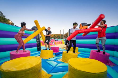 Four people play on a colorful inflatable arena, using foam sticks to compete while standing on large, round platforms.