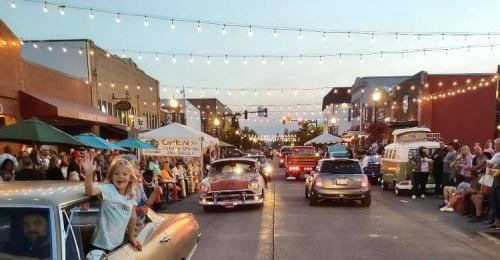 A lively street scene with classic cars, people enjoying the evening, and string lights illuminating the atmosphere.