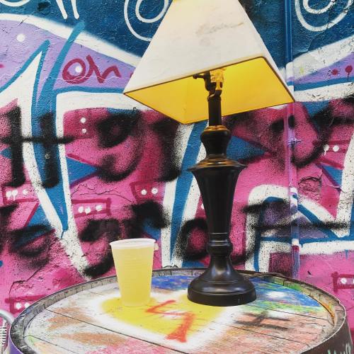 A lamp on a barrel next to a cup of yellow drink, set against a vibrant pink and blue graffiti wall.