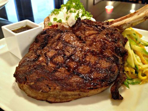 A grilled ribeye steak served with a side of pasta and a small dish of sauce, garnished with green onions.