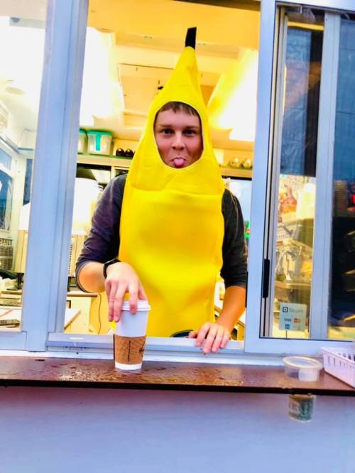 A person in a banana costume playfully sticks out their tongue while holding a coffee cup at a window.
