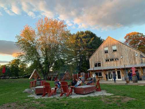 A cozy outdoor scene with a wooden building, fire pits, and people relaxing on red chairs under a colorful sky.