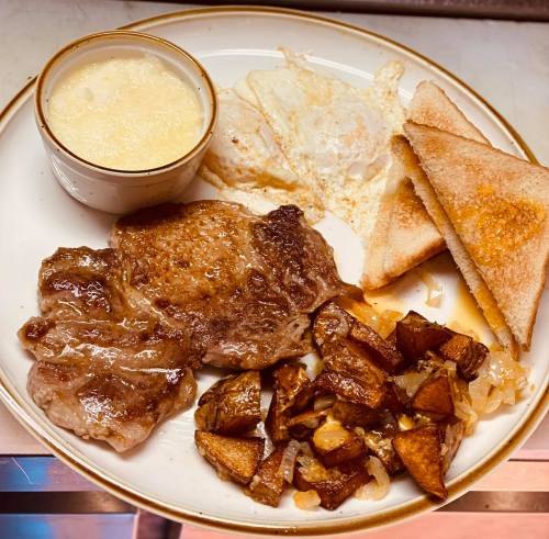 A plate with steak, fried eggs, toast, and diced potatoes with onions, accompanied by a small cup of sauce.