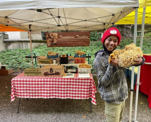 A smiling person in a red hat holds a large bundle of mushrooms at a market stall with various mushroom products.