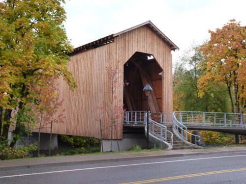 A wooden covered bridge surrounded by trees with autumn foliage, featuring a ramp and steps leading to it.
