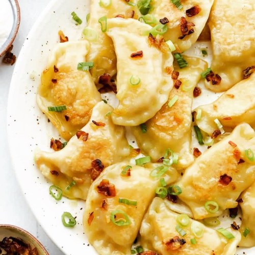 A plate of golden-brown dumplings topped with green onions and crispy bits, served with dipping sauce on the side.