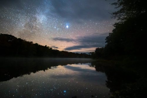 A serene night sky filled with stars reflects on a calm lake, surrounded by trees and a hint of mist.
