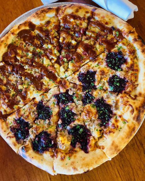 A pizza with various toppings, including brown sauce, herbs, and a mix of meats and vegetables, on a wooden table.