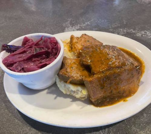 A plate of tender meat in sauce served with mashed potatoes and a side of red cabbage.