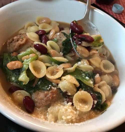 A bowl of soup with meatballs, assorted beans, spinach, and orecchiette pasta, garnished with cheese.