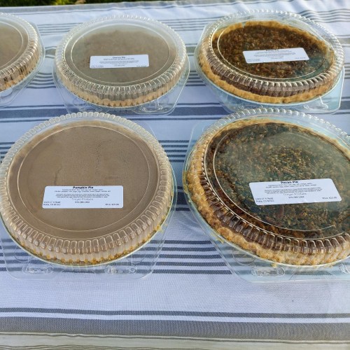 Five pies in clear plastic containers on a striped tablecloth, labeled with price tags.