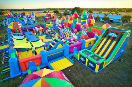 Aerial view of a colorful inflatable bounce park with slides, obstacles, and various play areas in a grassy field.