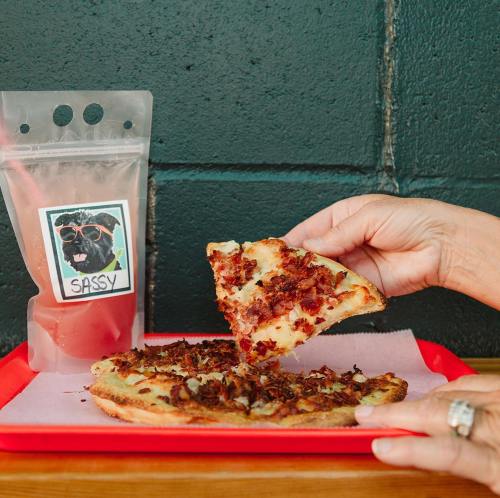 A hand holds a slice of pizza topped with bacon, next to a bag labeled "Sassy" on a red tray against a green wall.