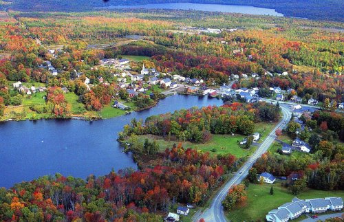 Aerial view of a colorful autumn landscape with a lake, homes, and vibrant foliage in various shades of red, orange, and green.