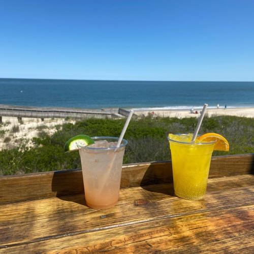 Two colorful drinks on a wooden table with a beach and ocean in the background under a clear blue sky.